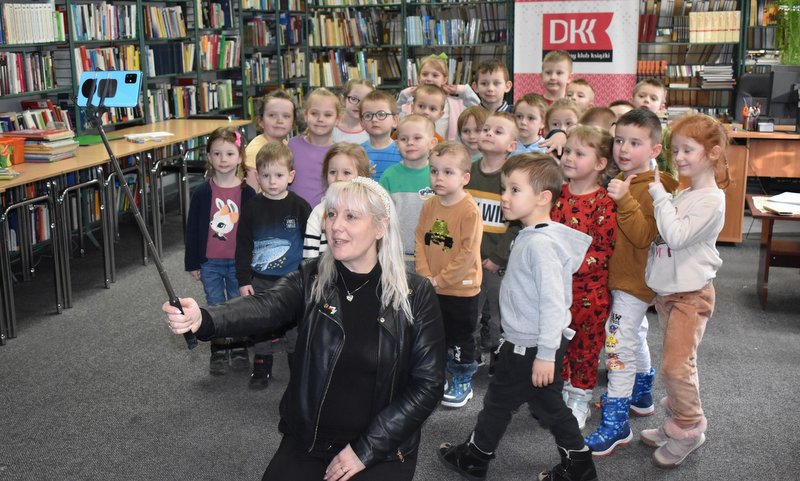 Dzieci w bibliotece pozujące do wspólnego zdjęcia z prowadzącą spotkanie