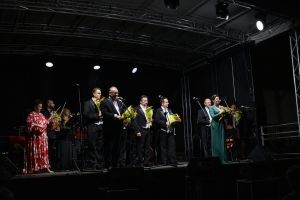 Zdjęcie przedstawia przemawiającą dyrektor BSCK , organizatora festiwalu oraz tenorów występujących podczas Gali Festiwalowej  XXVI Międzynarodowego Festiwalu im. Krystyny Jamroz. 
